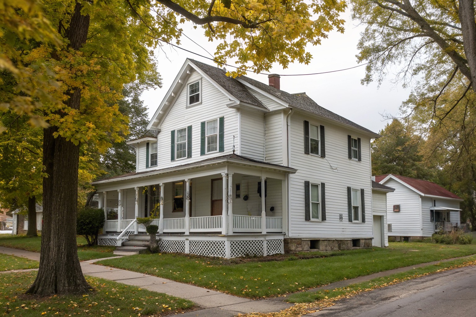 A 1920s white two-story frame house with a deep front porch and small basement windows on a maple-lined street in autumn