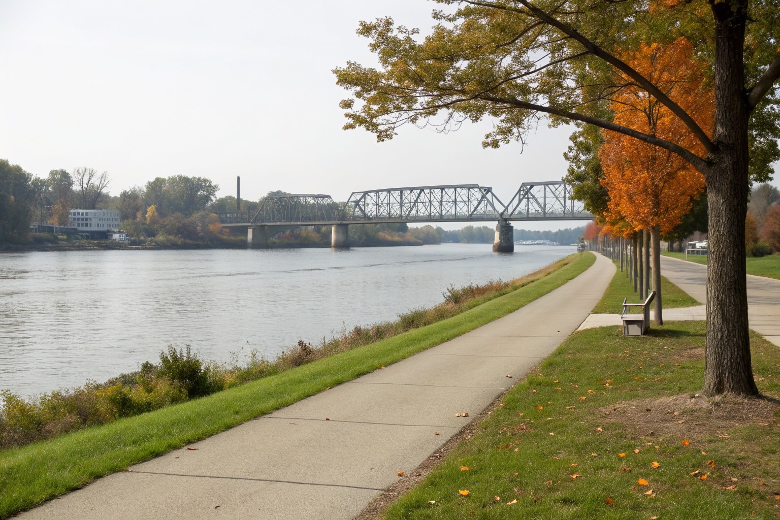 A paved riverside trail along a wide, calm river in early fall, with a steel bridge in the distance