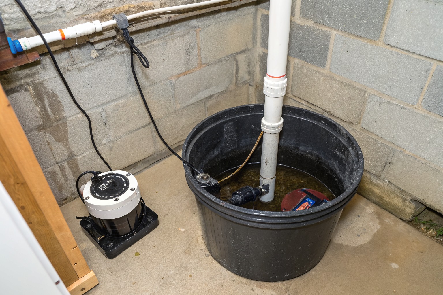 A black sump basin in a basement corner with a pump beside it and a white discharge pipe up the block wall